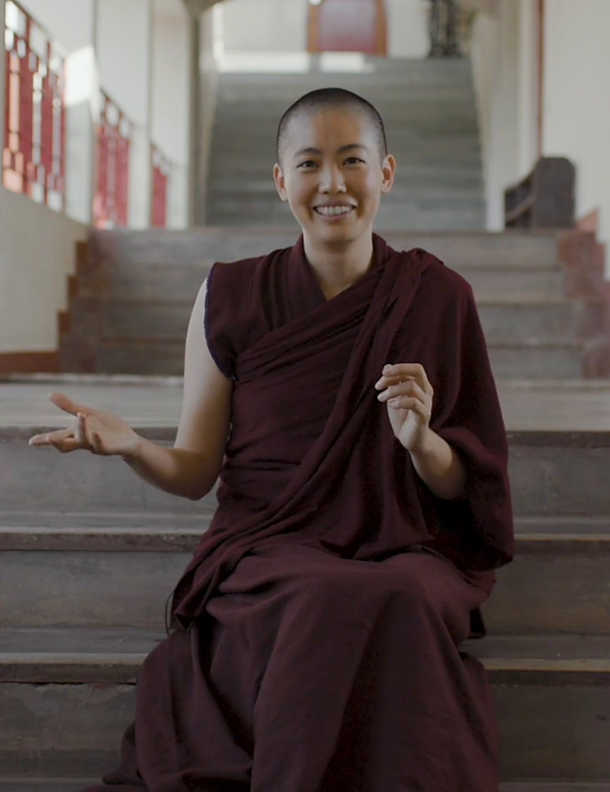 A Buddhist nun in maroon robes seated on temple steps, speaking, a Geshe-ma supported by The Maitri Project