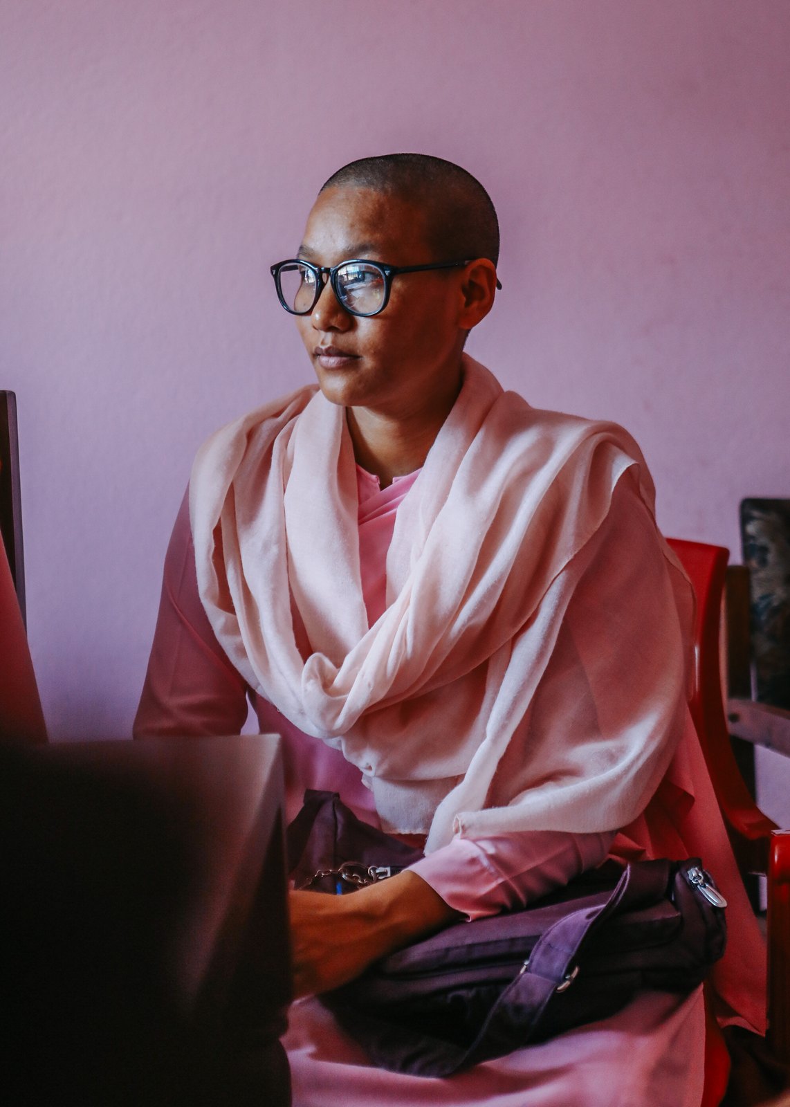 A nun in pink robes and glasses seated against a soft pink wall