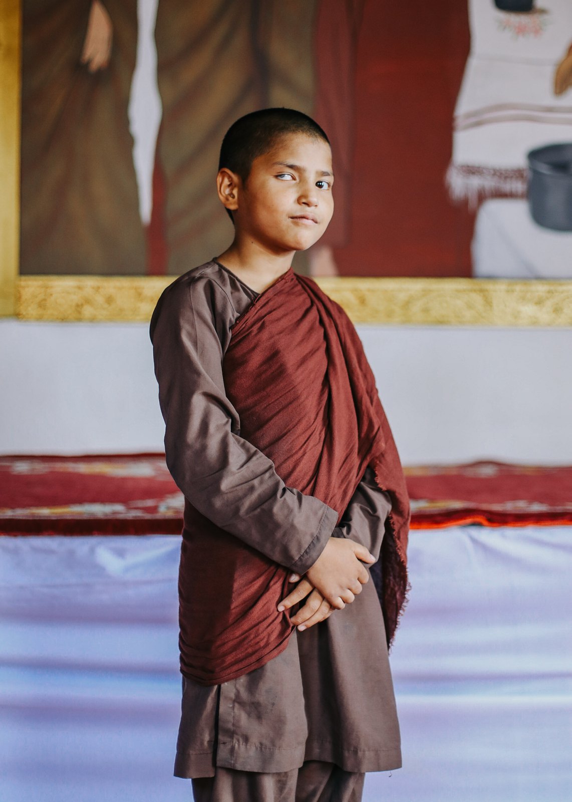 A young monk in maroon robes standing before a temple mural